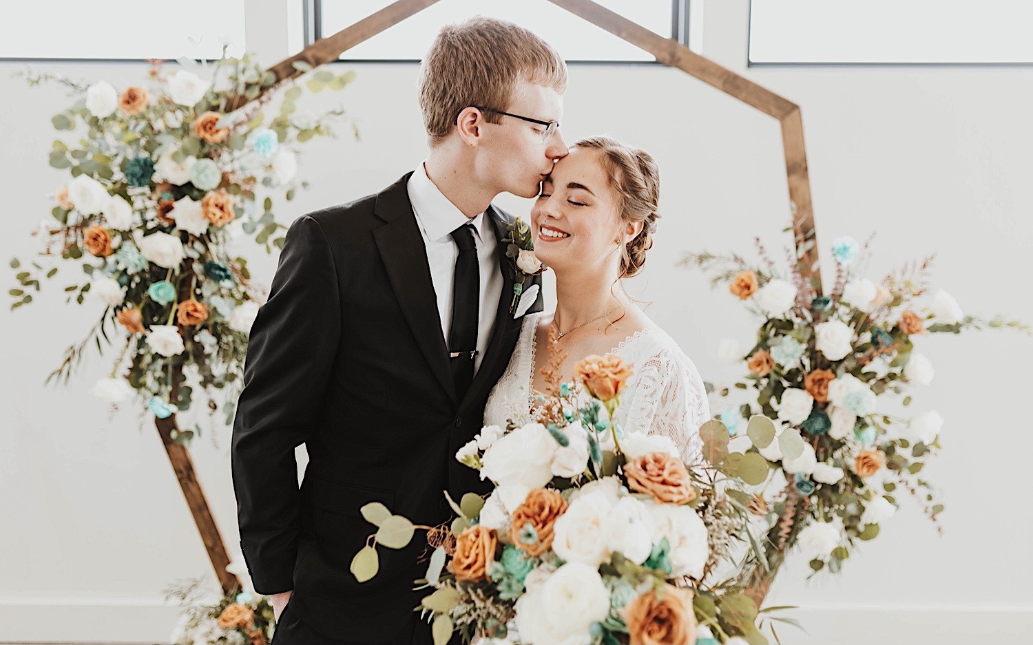 A bride and groom embrace on their wedding day in their ceremony space, the groom is kissing the bride on her temple A bride and groom embrace on their wedding day in their ceremony space, the groom is kissing the bride on her temple
