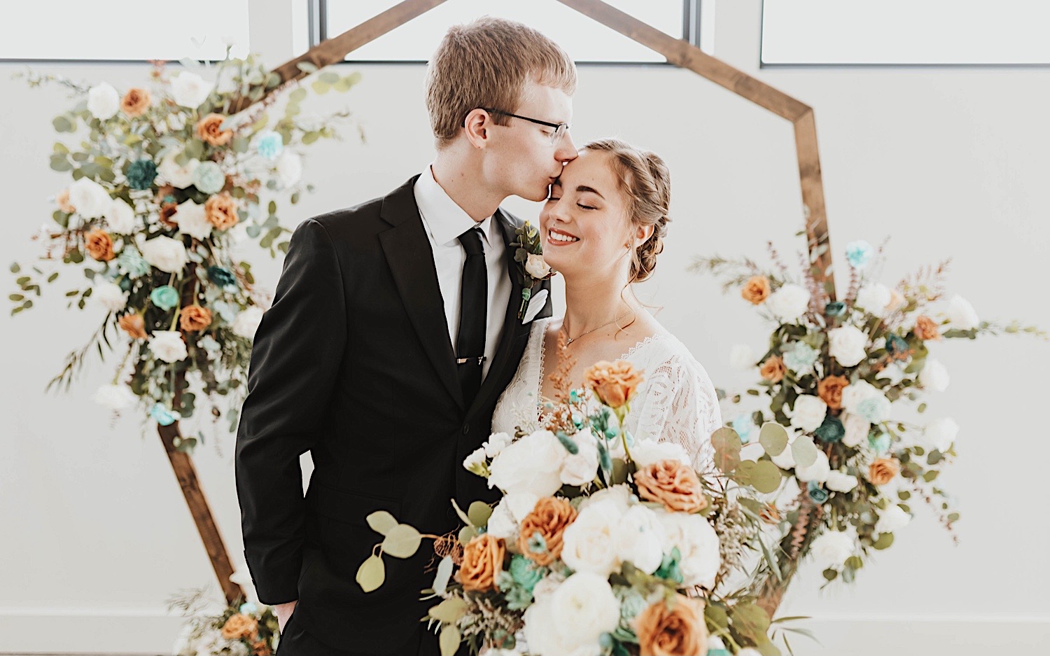 A bride and groom embrace on their wedding day in their ceremony space, the groom is kissing the bride on her temple