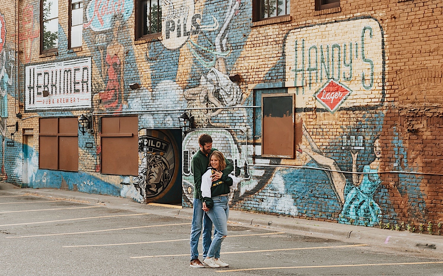 A man hugs a woman from behind while they stand in a parking lot in front of a mural on a brick wall during their engagement photos in the Twin Cities