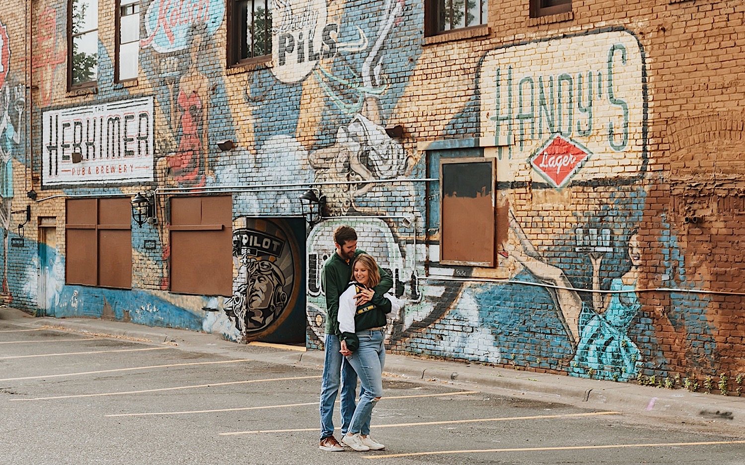 A man hugs a woman from behind while they stand in a parking lot in front of a mural on a brick wall during their engagement photos in the Twin Cities A man hugs a woman from behind while they stand in a parking lot in front of a mural on a brick wall during their engagement photos in the Twin Cities