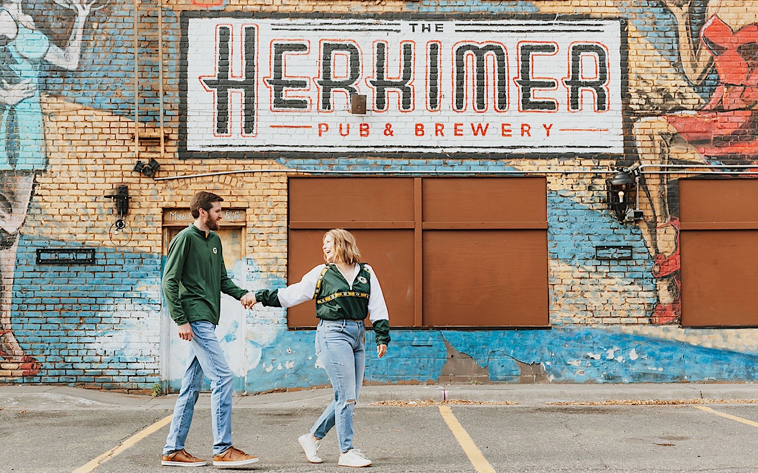 A couple walk hand in hand in front of a brick wall while looking at one another and smiling, photo taken by a Minneapolis Wedding Photographer A couple walk hand in hand in front of a brick wall while looking at one another and smiling, photo taken by a Minneapolis Wedding Photographer