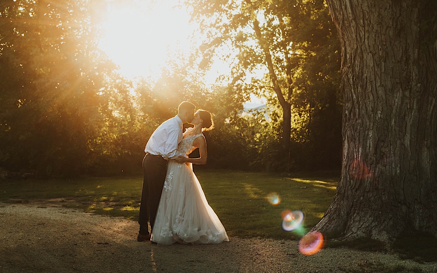 A bride and groom kiss one another in front of trees and bushes during sunset outside of their wedding venue Legacy Hill Farms