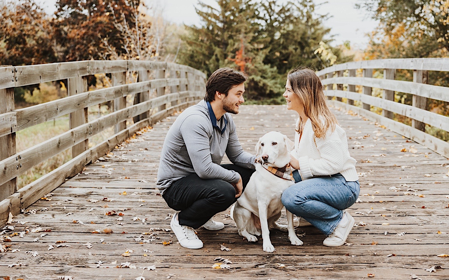 A man and woman crouch on a bridge and smile at one another while petting their dog in-between them during their fall engagement photos in the Twin Cities