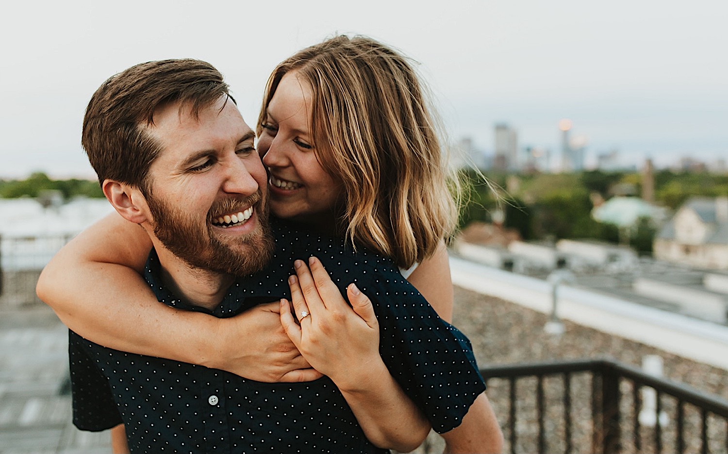 A man smiles as a woman rides on his back, the two are on the roof of their apartment in downtown Minneapolis and are taking engagement photos