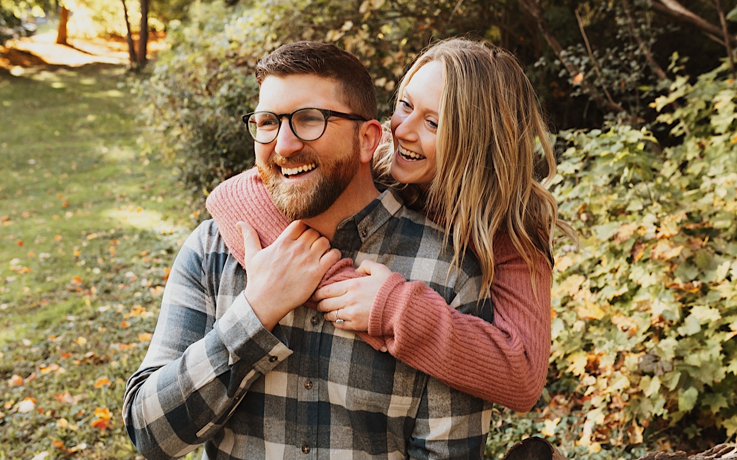 A man smiles as a woman hugs him from behind also smiling while the two are in a park with leaves on the ground during their fall engagement session in Winona A man smiles as a woman hugs him from behind also smiling while the two are in a park with leaves on the ground during their fall engagement session in Winona