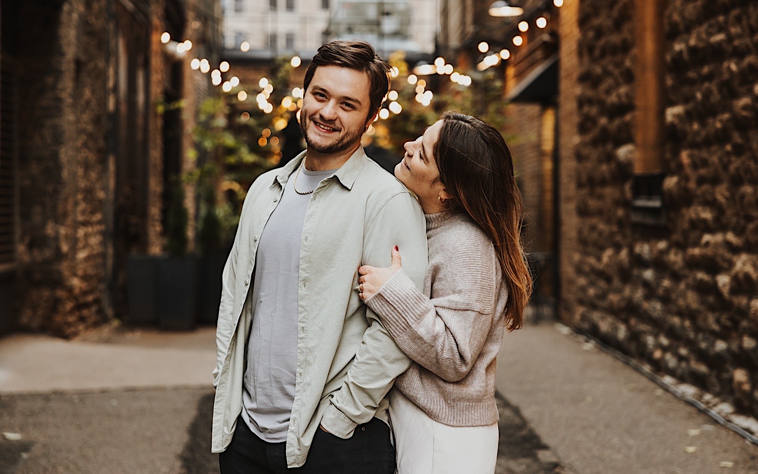 A man smiles at the camera while a woman hugs him from behind while taking engagement photos in the Twin Cities