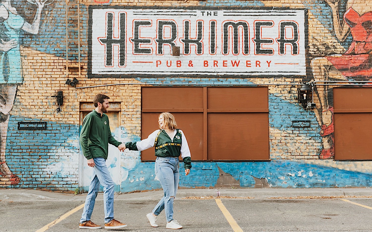 While taking engagement photos in downtown Minneapolis a woman looks back at a man while the two walk holding hands in front of a mural on a brick wall