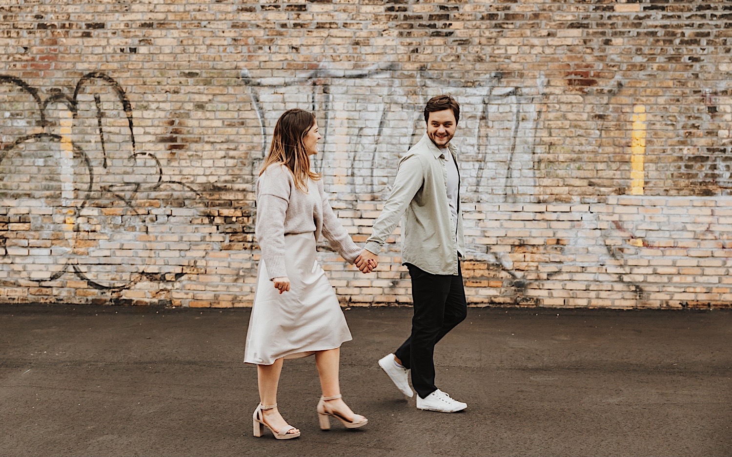 A man and woman walk holding hands with a brick wall behind them during their engagement photos in the Twin Cities