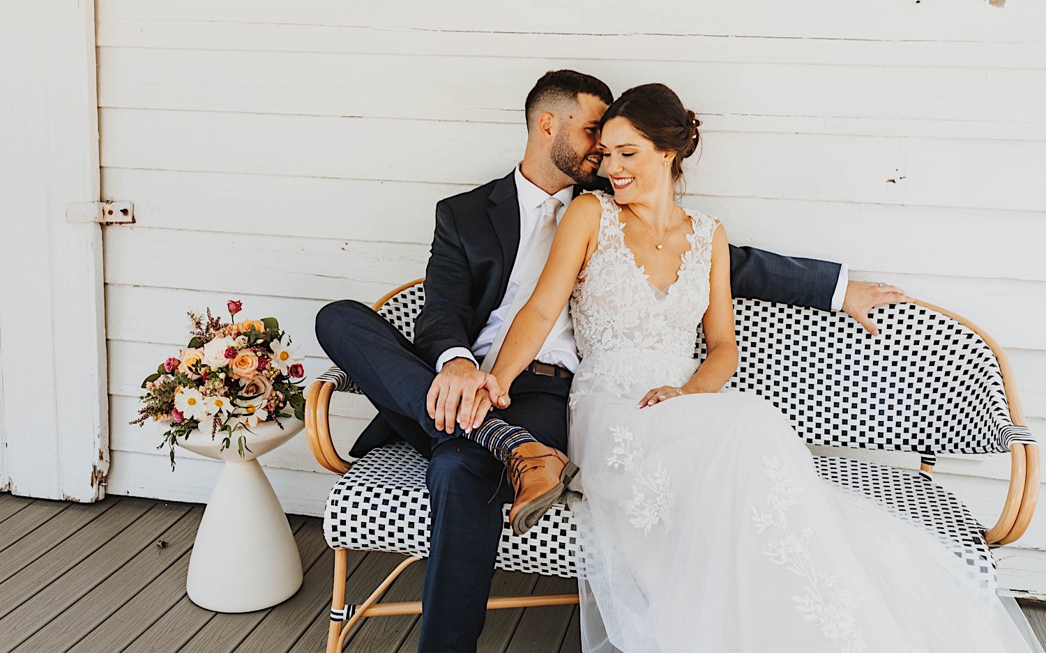 A bride and groom sit next to one another and smile on a bench outside of a house