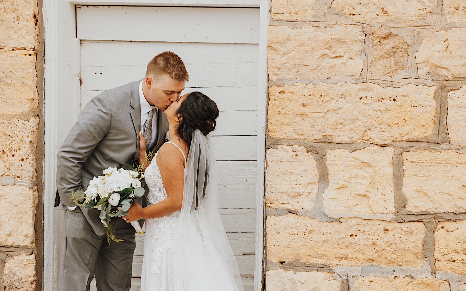 A bride and groom kiss in front of a white door on an old brick building, photo taken by a Minnesota Wedding Photographer
