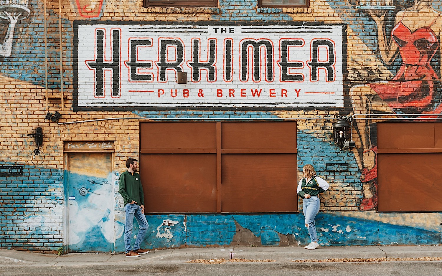 A man and woman look at one another while leaning against a brick wall with a mural for the Herkimer Brewery on it while taking engagement photos in downtown Minneapolis