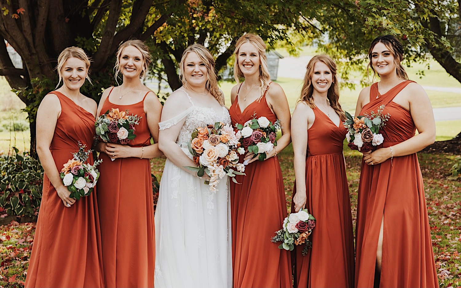 A bride and her bridesmaids stand together and smile at the camera while outdoors, photo taken by a Minneapolis wedding photographer