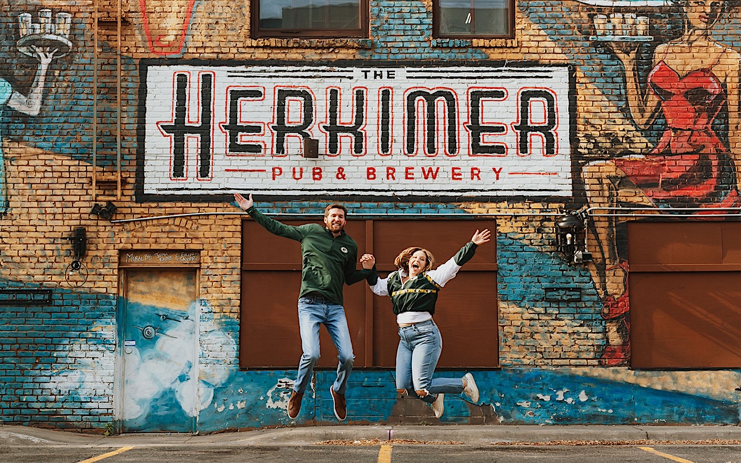 A couple hold hands and jump in the air while smiling at the camera, behind them is a brick wall with a mural on it for the Herkimer Brewery