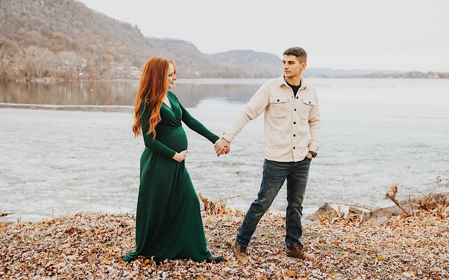 A couple hold hands and look at one another while standing in front of a lake, the woman holds her belly to show off her baby bump, photo taken by a Minneapolis wedding photographer