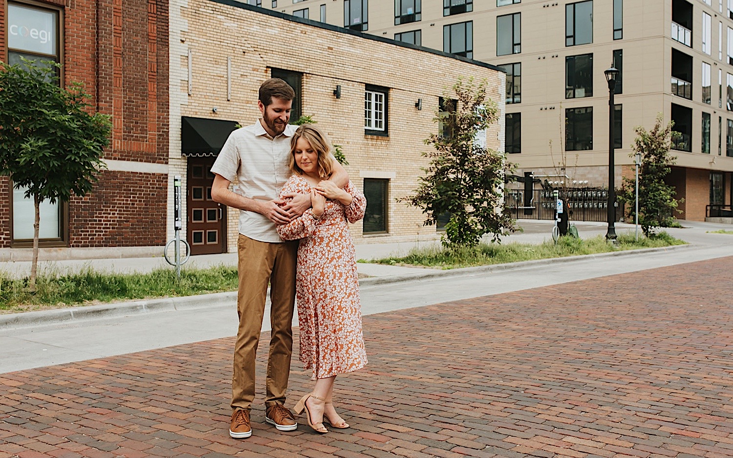 A man hugs a woman from behind while they stand on a brick road together in the Twin Cities while taking engagement photos A man hugs a woman from behind while they stand on a brick road together in the Twin Cities while taking engagement photos