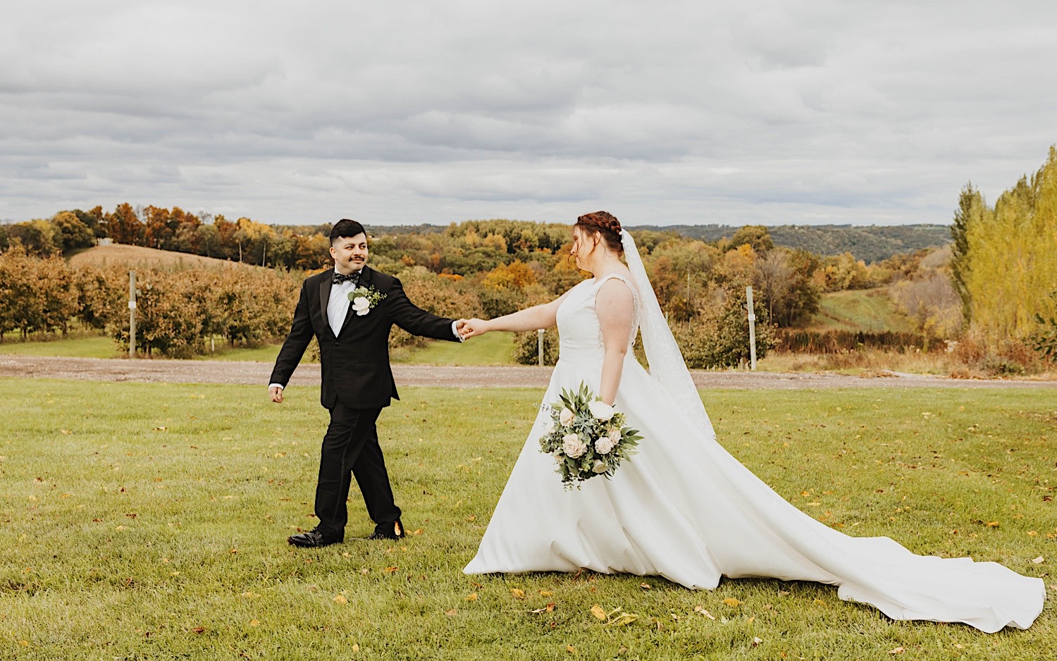 A bride and groom hold hands while walking through a field together on a cloudy day