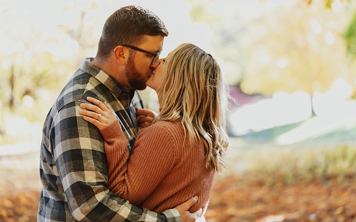 A man and woman kiss one another and embrace as the woman places her hand on the man's shoulder showing her engagement ring, leaves are on the ground around them A man and woman kiss one another and embrace as the woman places her hand on the man's shoulder showing her engagement ring, leaves are on the ground around them