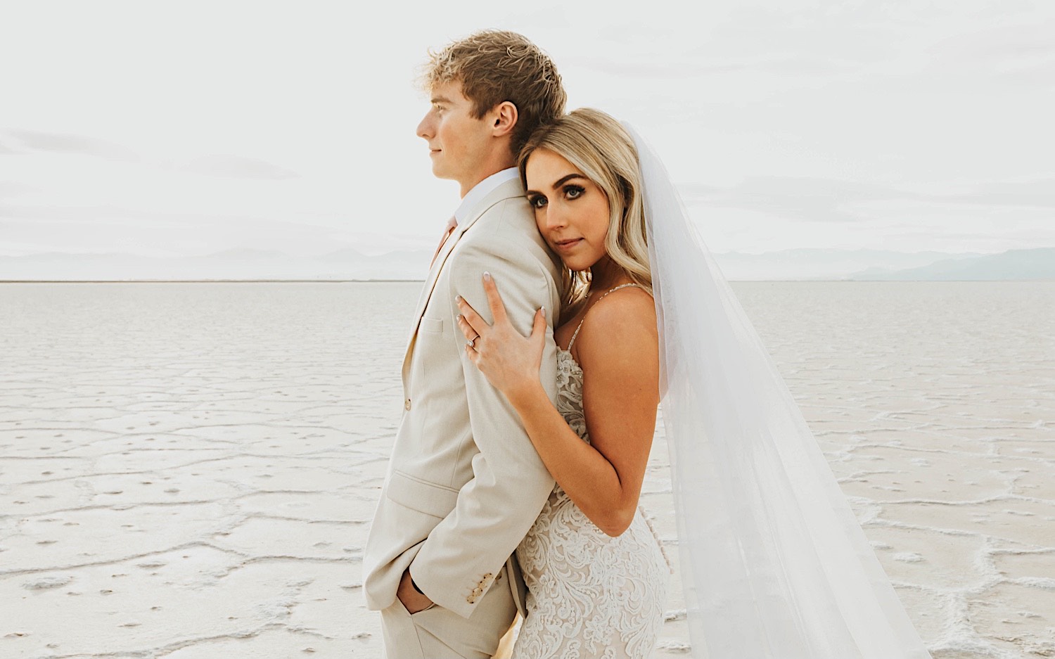 A groom looks off camera as the bride embraces him from behind and looks towards the camera while standing in the Utah Salt Flats on the day of their elopement A groom looks off camera as the bride embraces him from behind and looks towards the camera while standing in the Utah Salt Flats on the day of their elopement