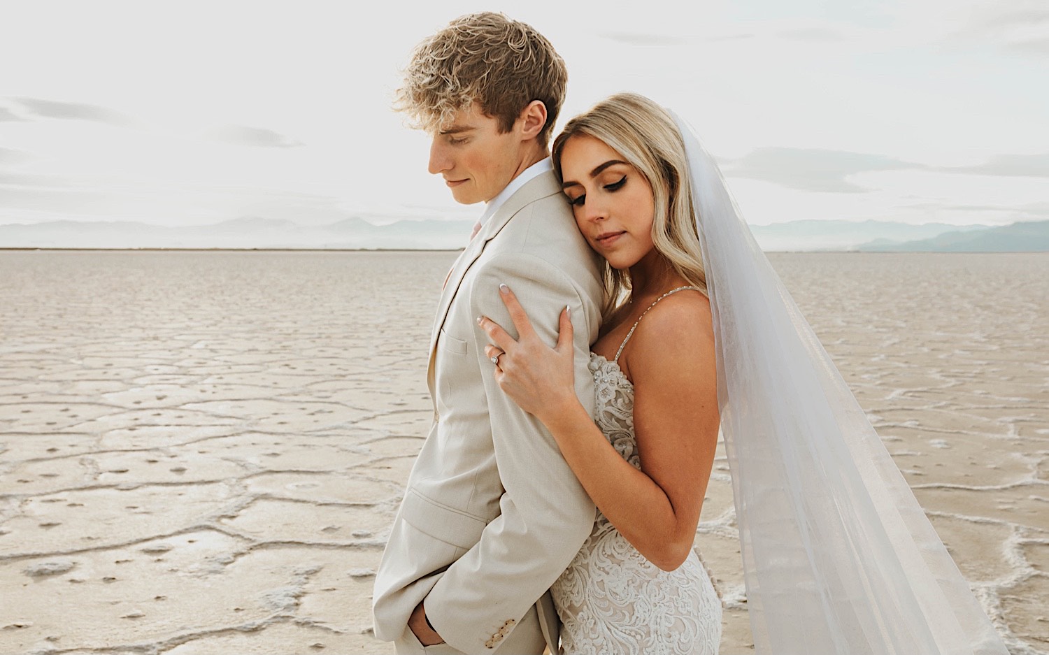A bride stands behind the groom during their elopement in the Utah Salt Flats and rests her head on his shoulder as they embrace A bride stands behind the groom during their elopement in the Utah Salt Flats and rests her head on his shoulder as they embrace