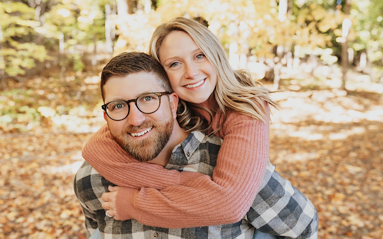 Portrait photo of a couple on a trail covered in leaves where the woman is riding on the man's back as they both smile at the camera Portrait photo of a couple on a trail covered in leaves where the woman is riding on the man's back as they both smile at the camera