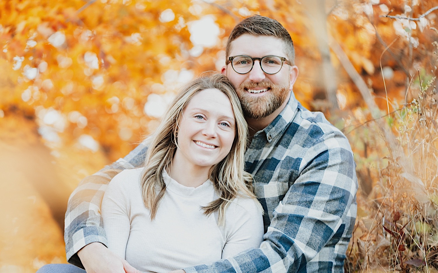 A couple sit with one another and smile at the camera in front of fall leaves on a tree during their engagement session in Minnesota