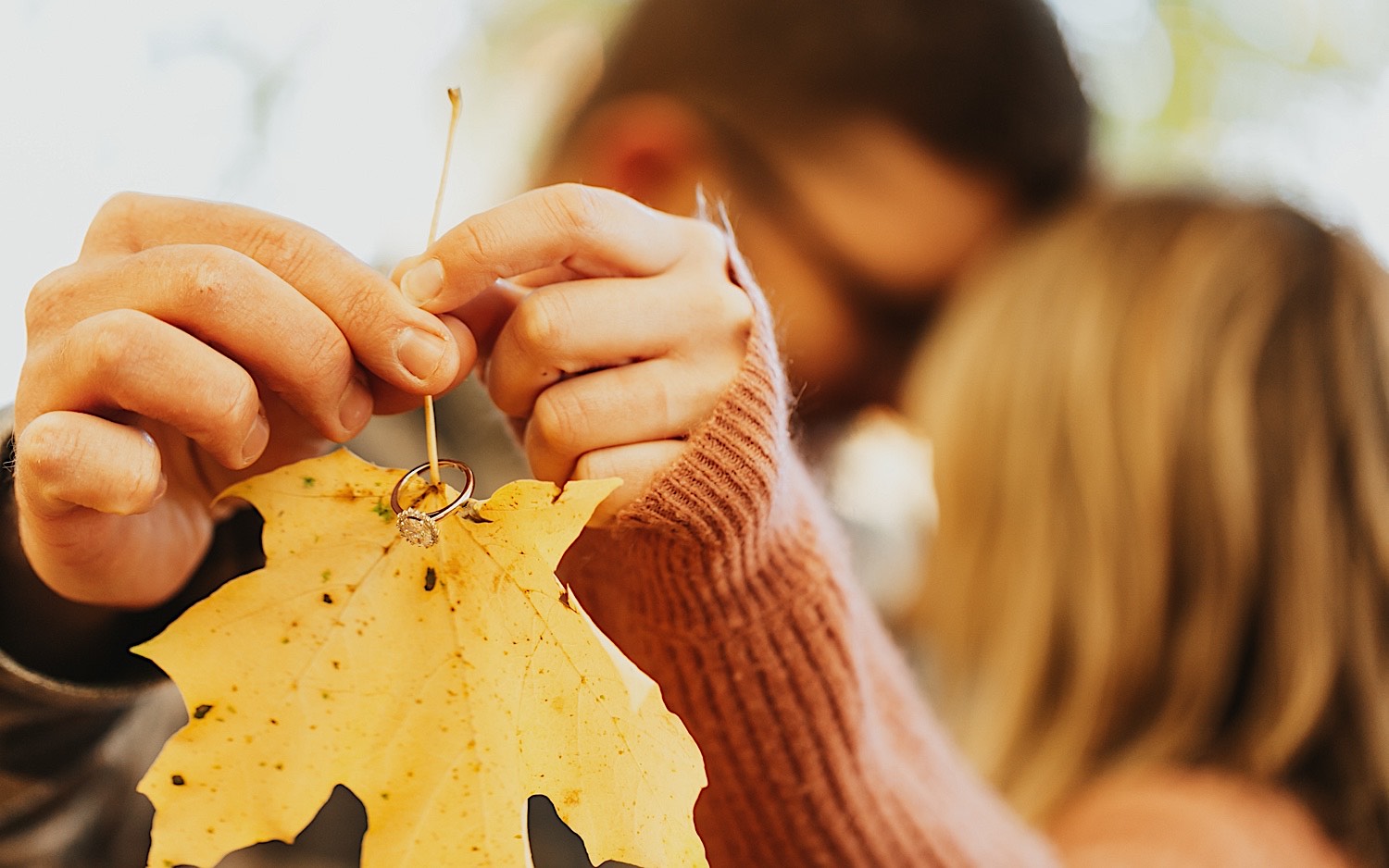 Detail photo taken during a fall engagement session in Winona of a yellow leaf being held by a couple who are kissing one another in the background, on the leaf's stem is an engagement ring Detail photo taken during a fall engagement session in Winona of a yellow leaf being held by a couple who are kissing one another in the background, on the leaf's stem is an engagement ring