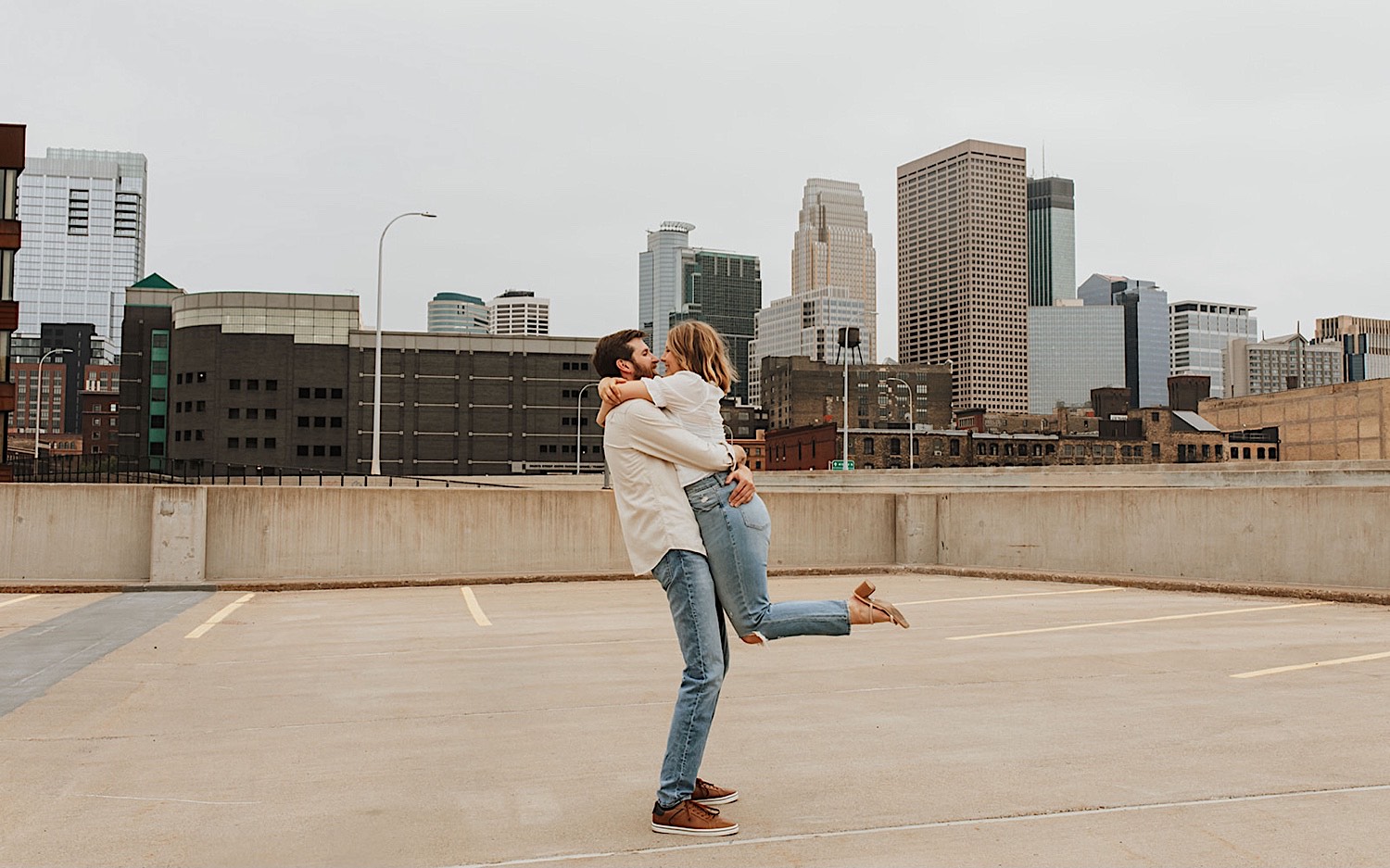 A couple smile at one another while the man lifts the woman in the air as the two have their engagement photos taken while on a parking garage in downtown Minneapolis