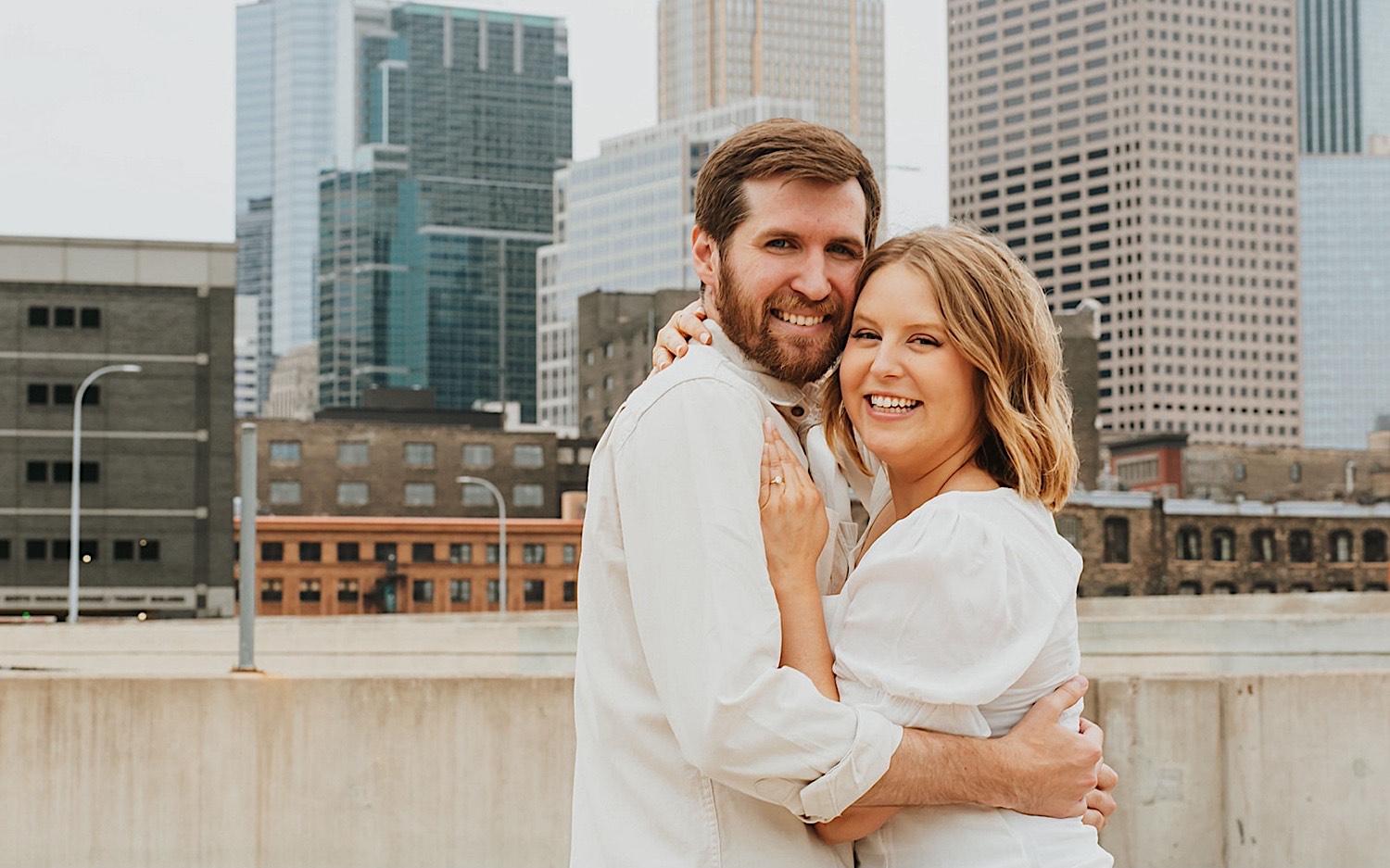 A couple embrace and smile at the camera while the woman shows off her engagement ring as they get their engagement photos taken while on the top level of a parking garage in downtown Minneapolis