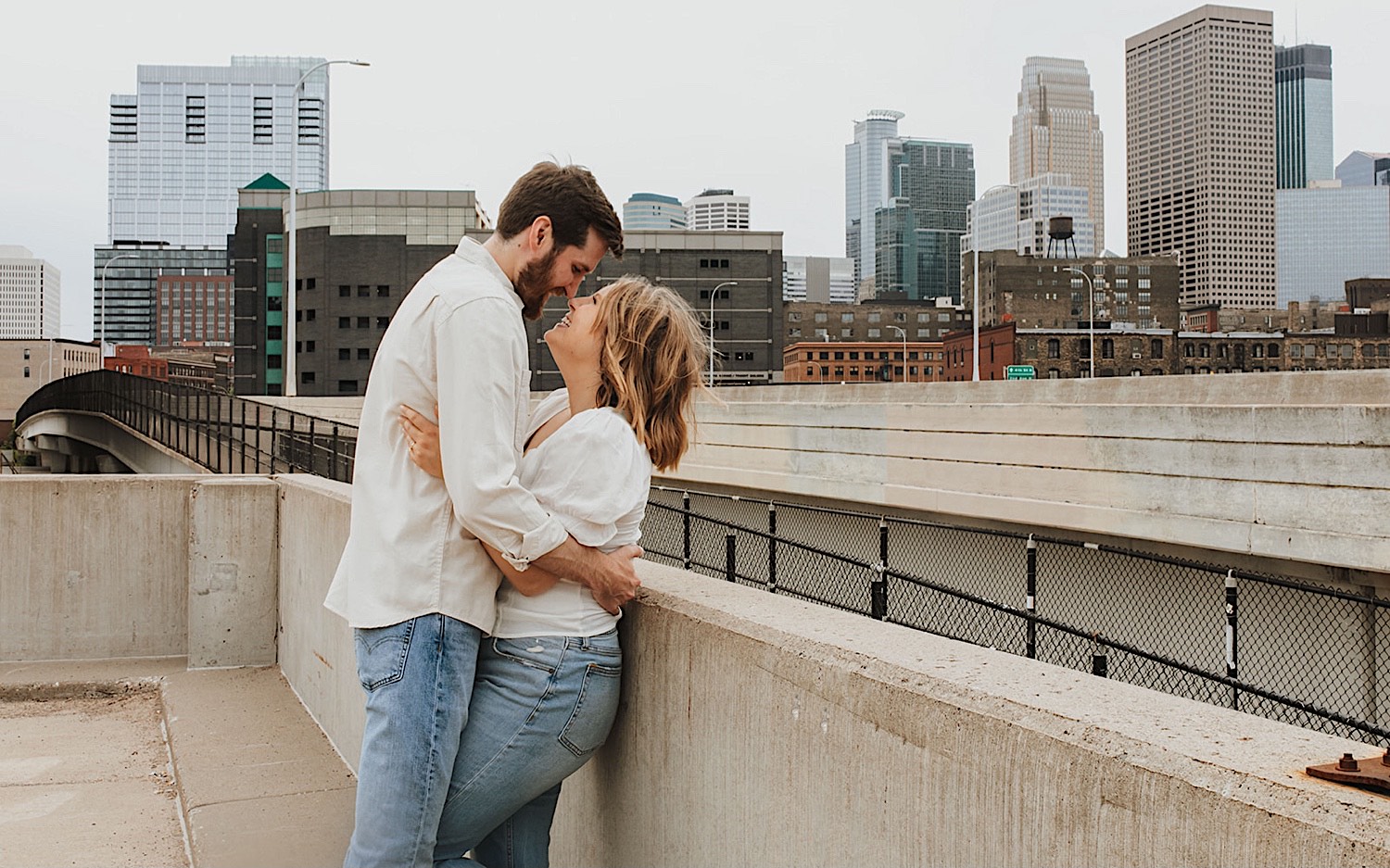 A couple smile at one another while the woman leans against a concrete wall as the two have their engagement photos taken on the top level of a parking garage in downtown Minneapolis