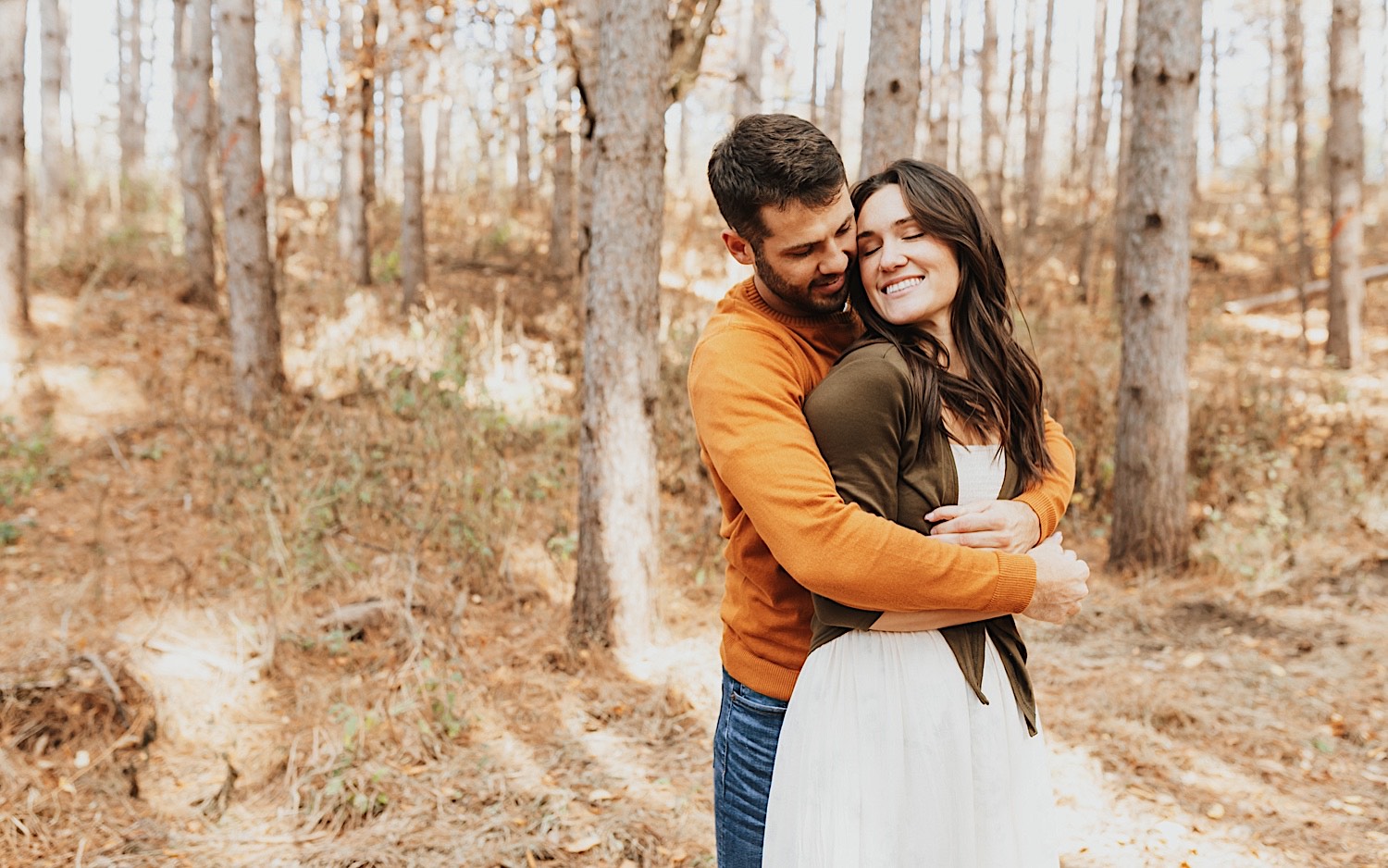 A woman smiles with her eyes closed as a man hugs her from behind while they stand in a forest with fall leaves on the ground during their engagement session in Minnesota