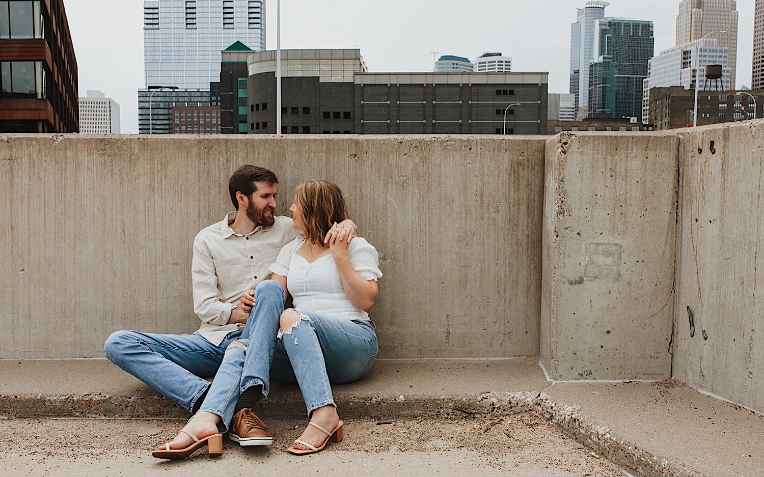 While having their engagement photos taken a couple sit next to one another and smile at each other while on the top level of a parking garage in downtown Minneapolis