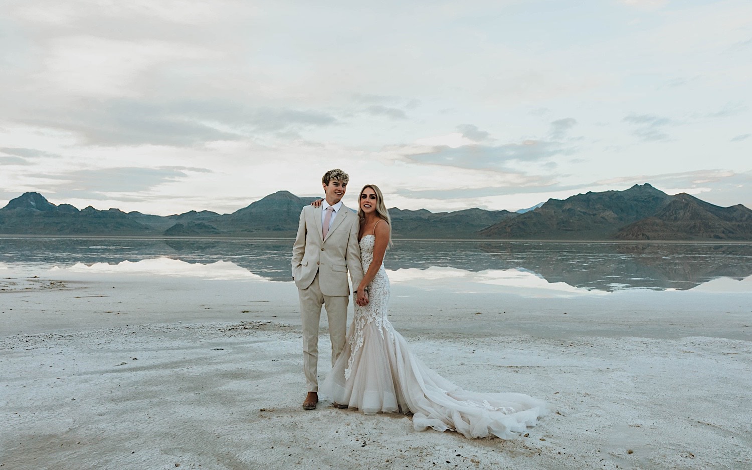 A bride and groom stand next to one another and hold hands while smiling at the camera standing in the Utah Salt Flats on their elopement day with the mountains behind them A bride and groom stand next to one another and hold hands while smiling at the camera standing in the Utah Salt Flats on their elopement day with the mountains behind them