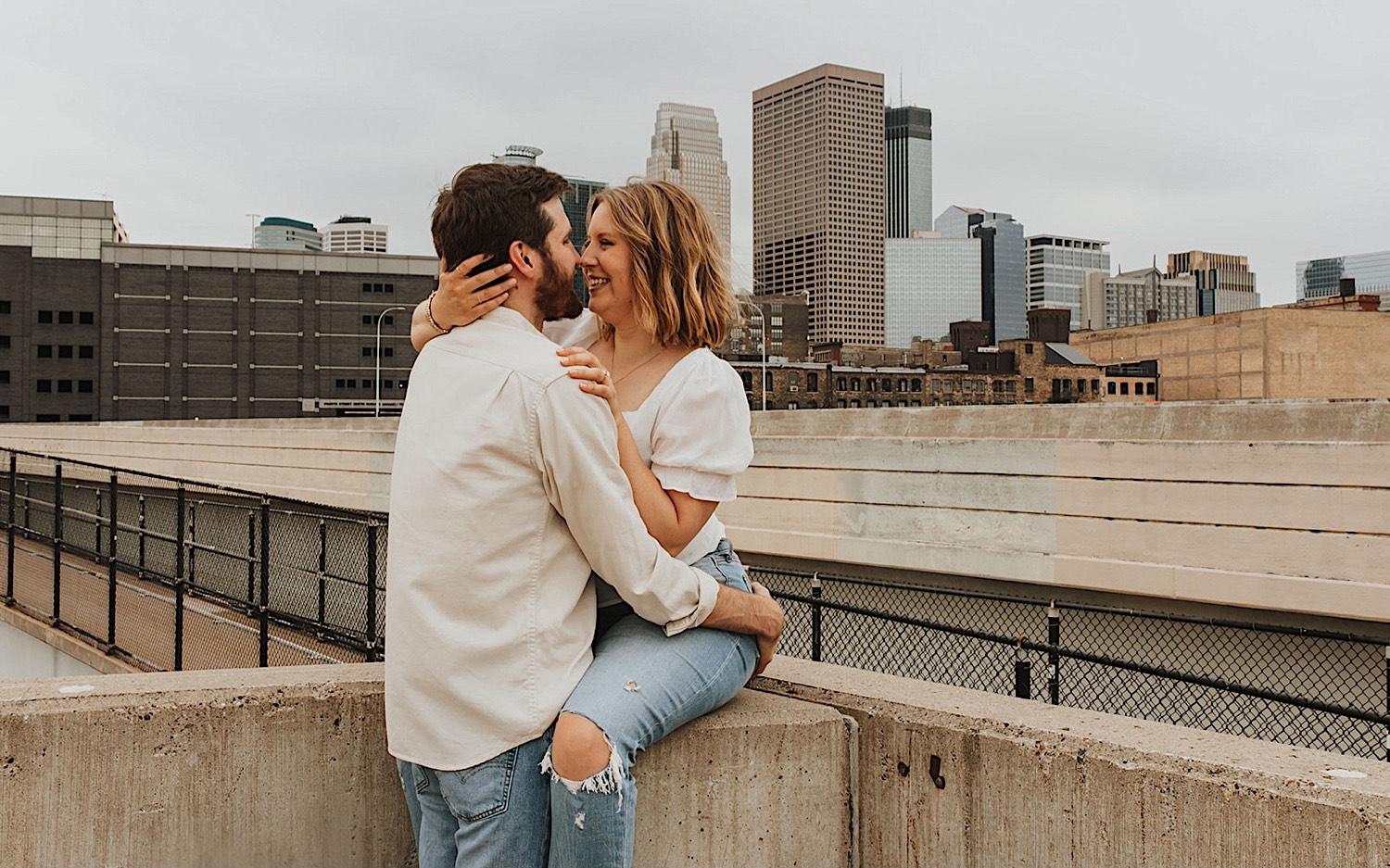 A woman sits on a ledge of a parking garage in downtown Minneapolis and smiles as a man hugs her and is about to kiss her while the two are having their engagement photos taken