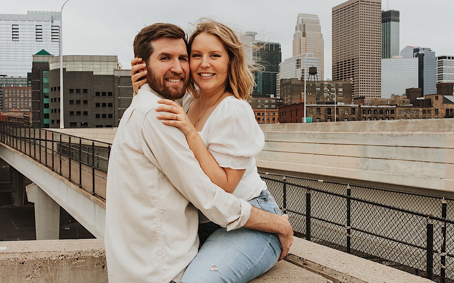 A woman sits on the ledge of a parking garage in downtown Minneapolis and smiles at the camera as a man is hugging her and also smiling at the camera while the two have their engagement photos taken