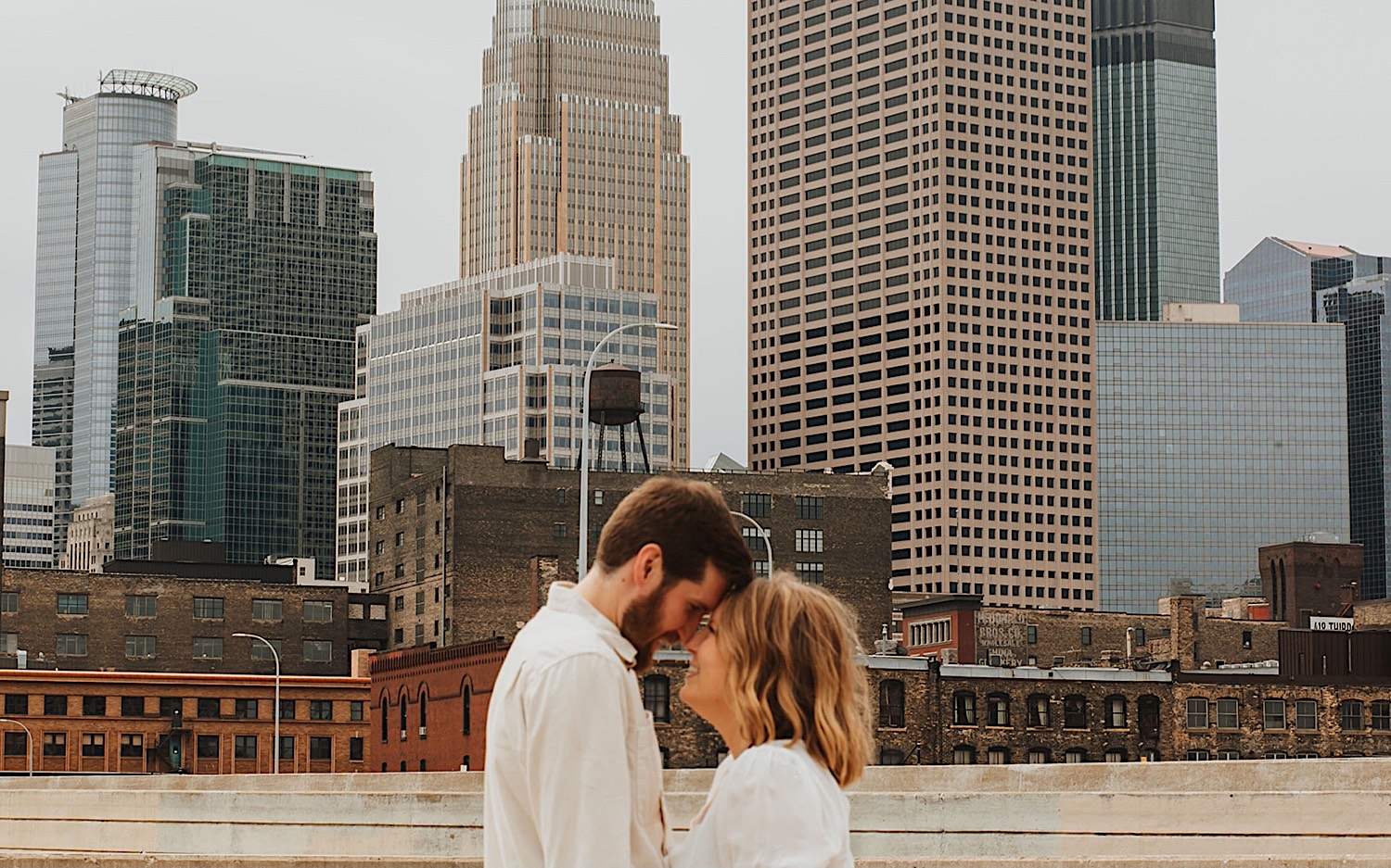 A couple touch foreheads together while taking engagement photos with the skyline of downtown Minneapolis behind them