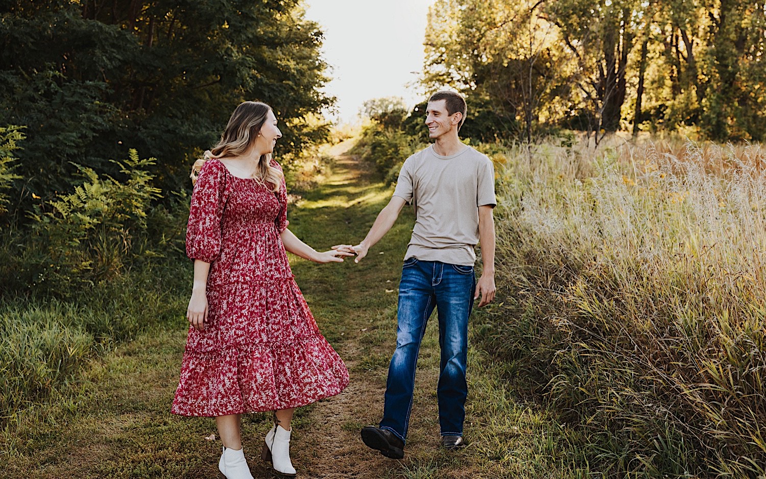 A couple on a grass path between a field and a forest smile at one another and hold hands