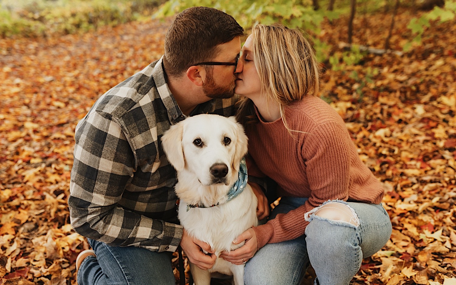 A couple kiss one another with fall colored leaves on the ground around them during their engagement session in Minnesota, between them is their dog who is looking at the camera