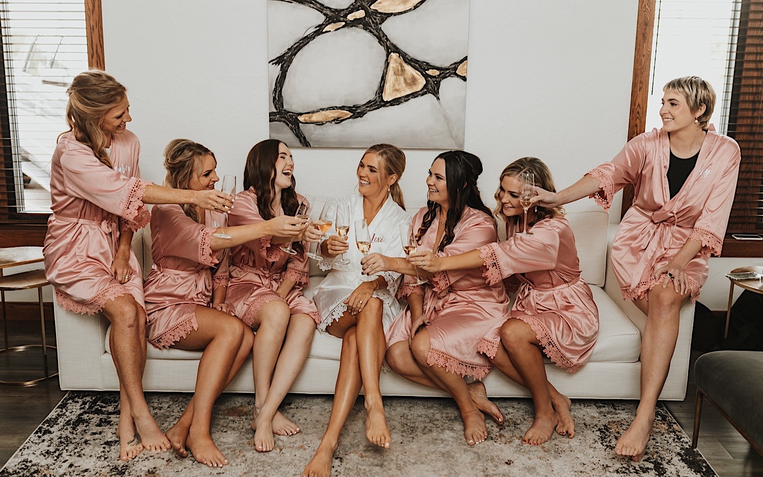 A bride and her bridesmaids sit on a couch and drink champagne with one another before their wedding day
