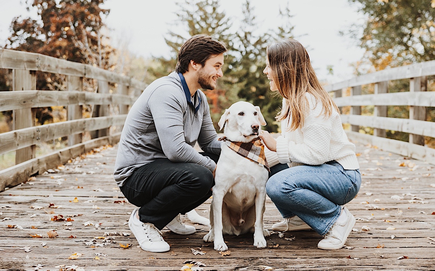 A couple crouch down next to their dog while smiling at one another during their engagement session at a park in Minneapolis