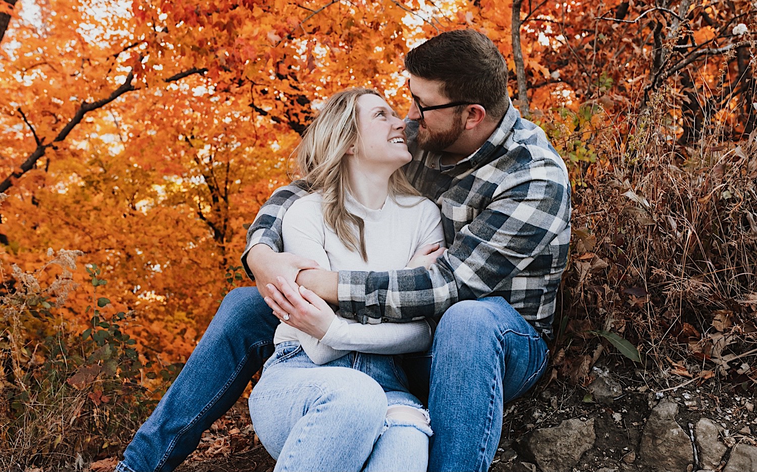 A woman sits between a man's legs and looks up into his eyes as he looks into hers, around them are orange leaves during their Fall engagement session in Winona
