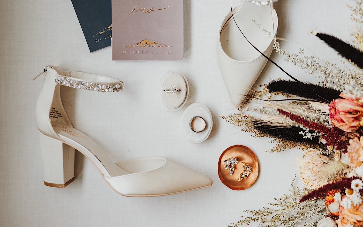 A wedding day flatlay consisting of shoes, vows, earrings, wedding rings, and a bouquet