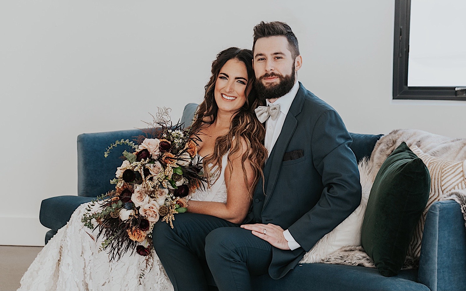 A bride and groom smile at the camera while sitting next to one another on a couch inside of Woodhaven Weddings + Events