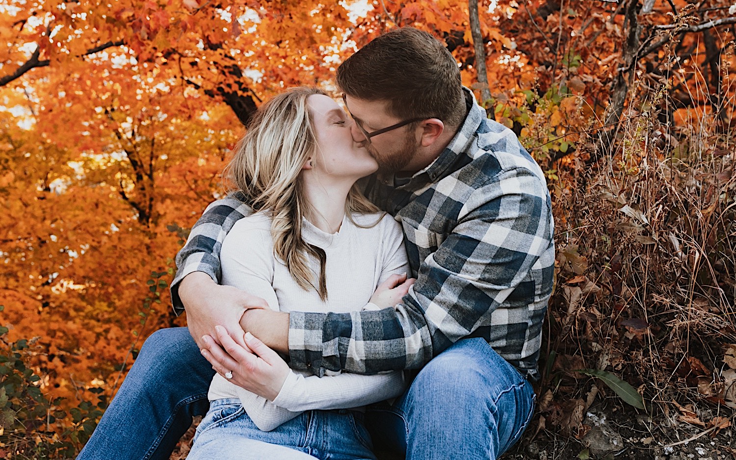 A couple sitting on the ground kiss as the man wraps his arms around the woman, around them are orange leaves on trees during their Fall engagement session in Winona