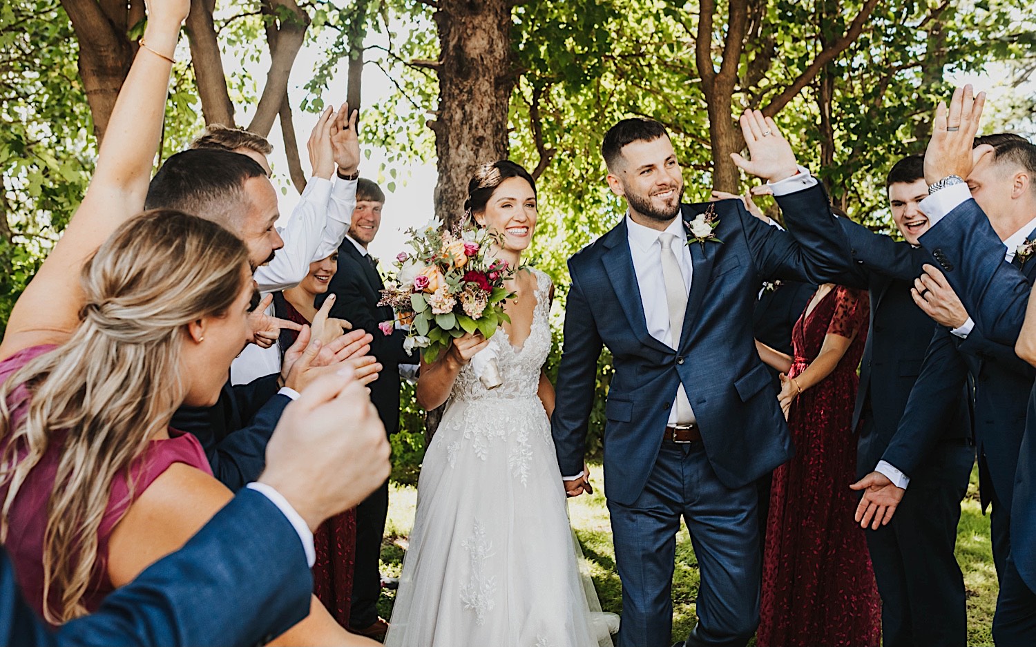 A bride and groom walk hand in hand and smile as their wedding parties cheer for them on either side