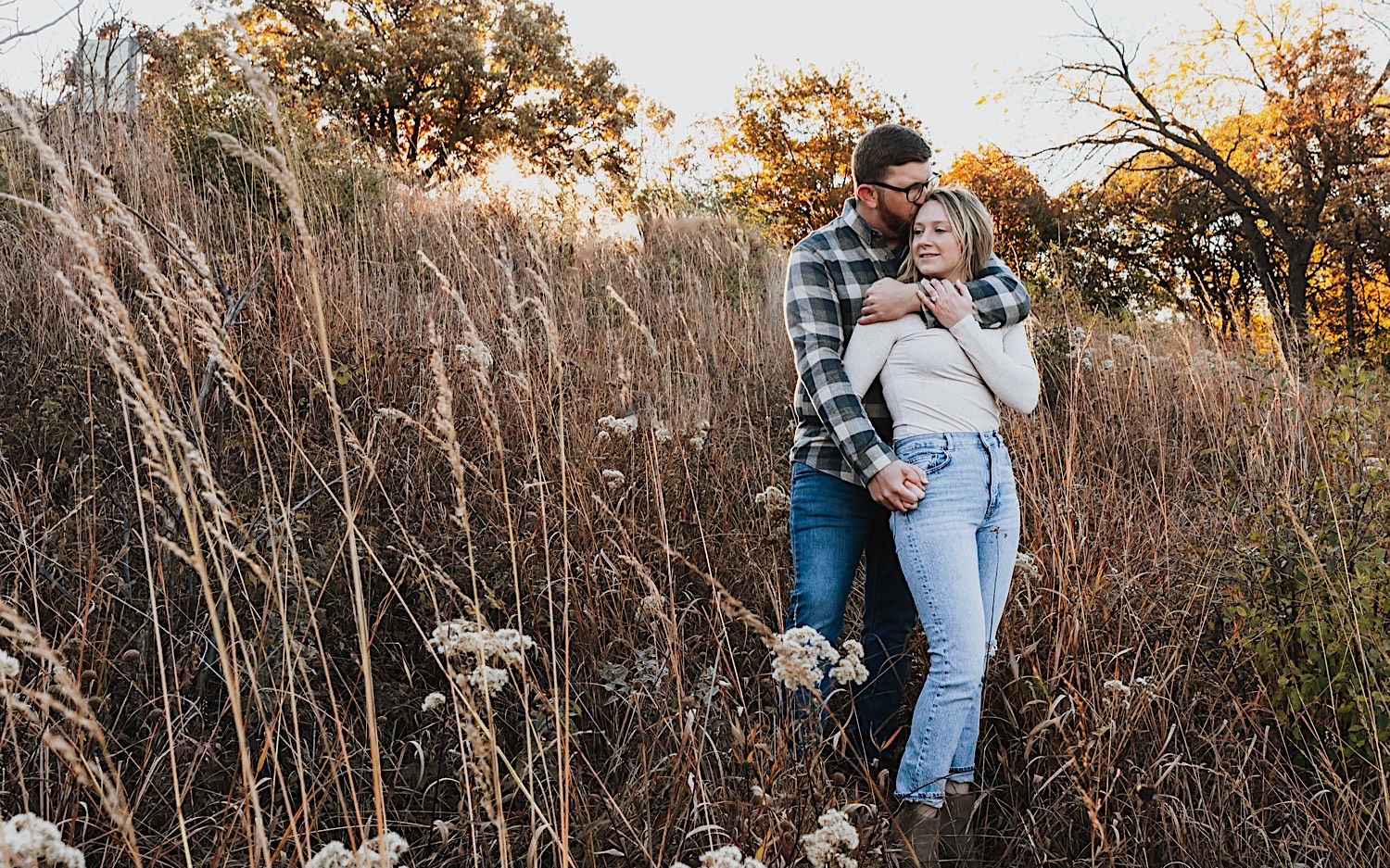A woman looks over her shoulder as a man hugs her from behind and kisses her temple while they stand in a field with fall leaves behind them during their engagement session in Winona