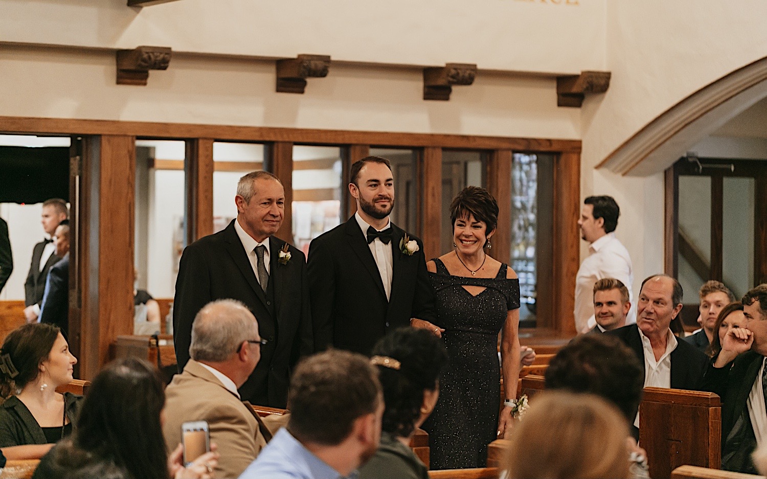 A groom is walked down the aisle by his mother and father