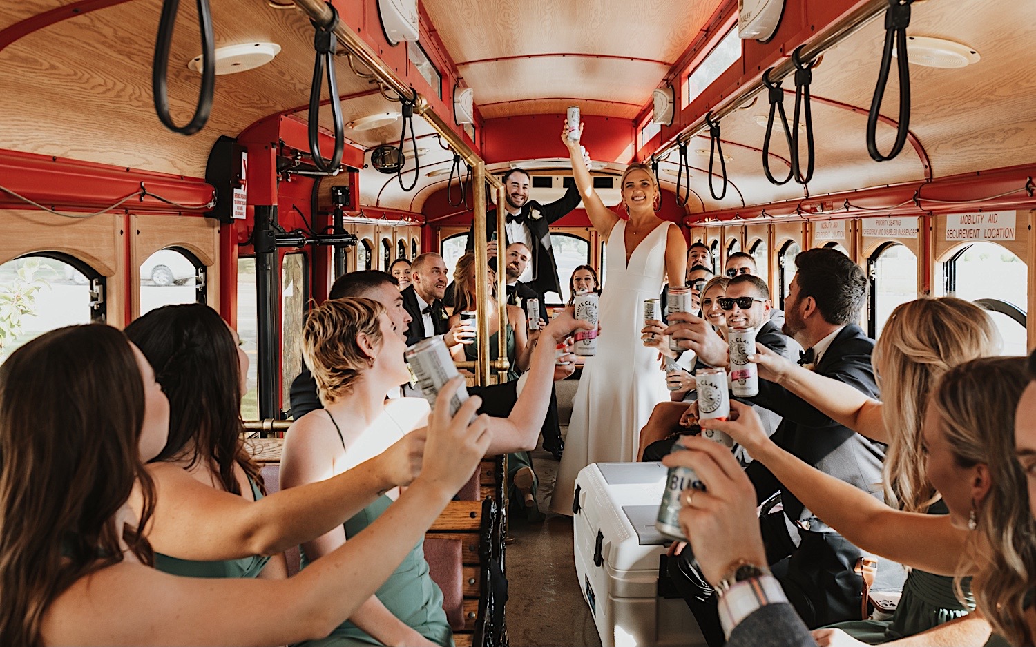 A bride and groom ride a trolley with their wedding parties while all raising a white claw for a toast