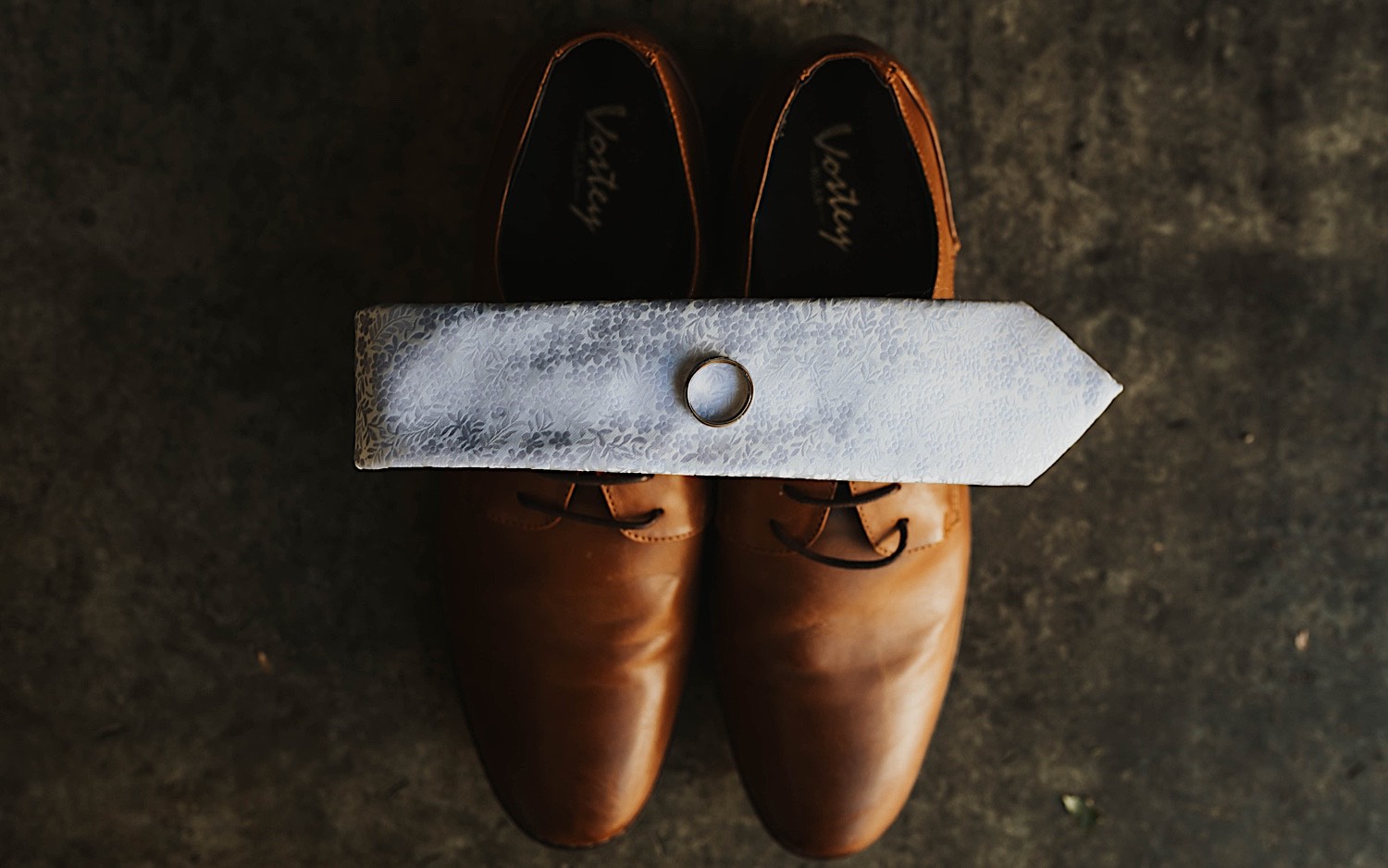 Top down photo of a wedding ring atop a folded tie which is resting on a pair of men's dress shoes