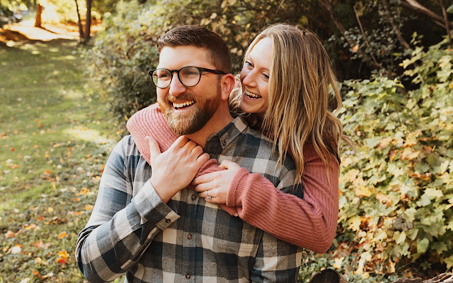 A man smiles as a woman hugs him from behind also smiling while the two are in a park with leaves on the ground during their fall engagement session in Winona