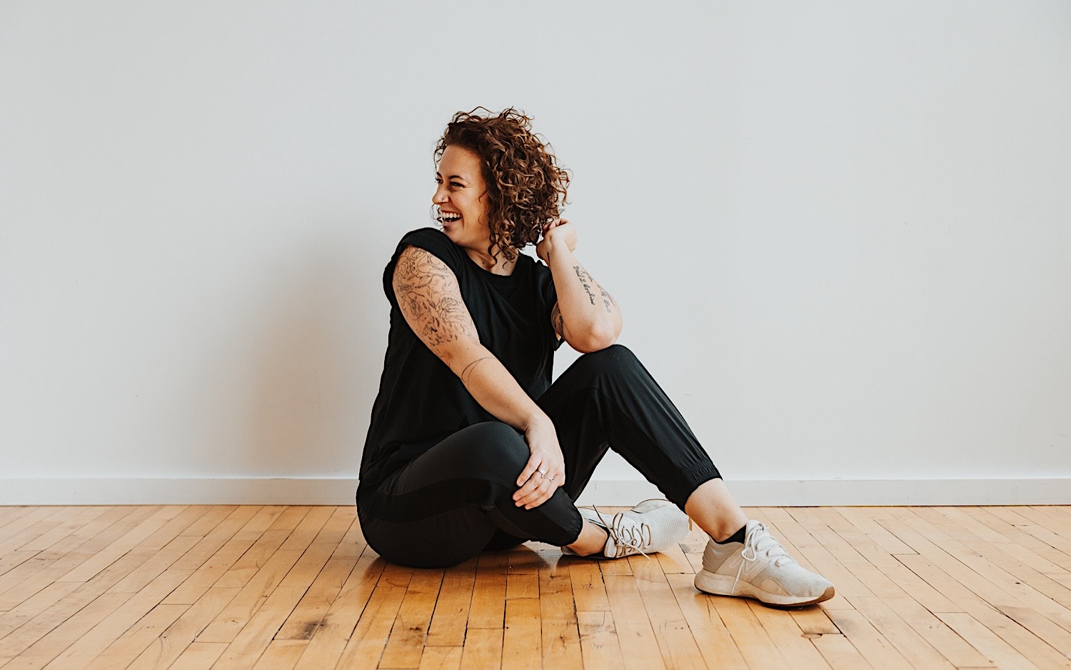 The Minnesota Wedding Photographer Chelsea of Sage River Studios sits on a wood floor and smiles while playing with her hair